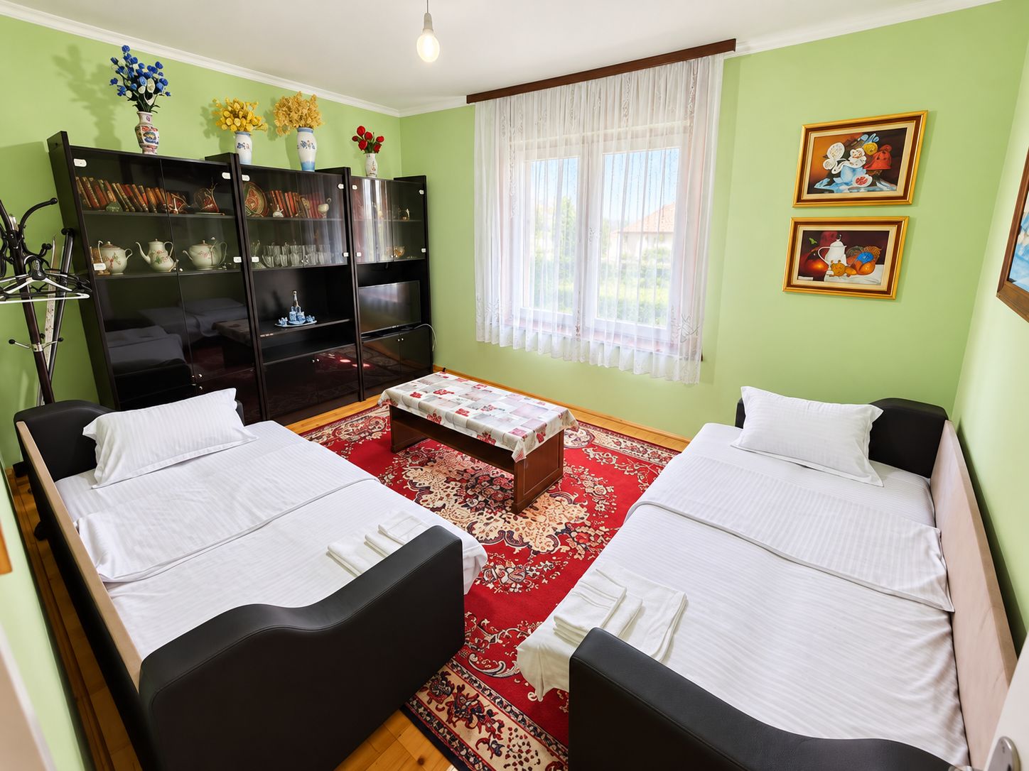 A room with two beds, a bookshelf, a window with curtains, and two framed pictures on the wall.