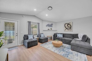 Ein modernes Wohnzimmer mit einem grauen Ecksofa, zwei Sesseln, einem Couchtisch und einem gemusterten Teppich.