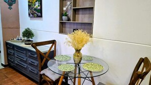 A dining area with a round glass table, four chairs, a vase with dried flowers, a wall-mounted shelf with decorative items, and a painting.