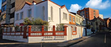 A street view with a row of buildings, including a white building with a red and white fence, and a brown building with a red roof.