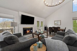 Un salon avec une cheminée, un téléviseur fixé au mur, un coin repas avec des chaises et une table, et une grande fenêtre.