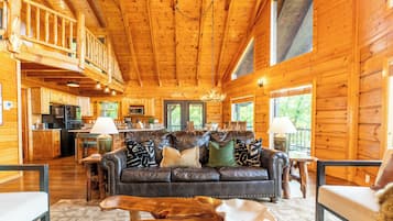 A log cabin interior with a living room, kitchen, and dining area, featuring wooden walls and a high ceiling.