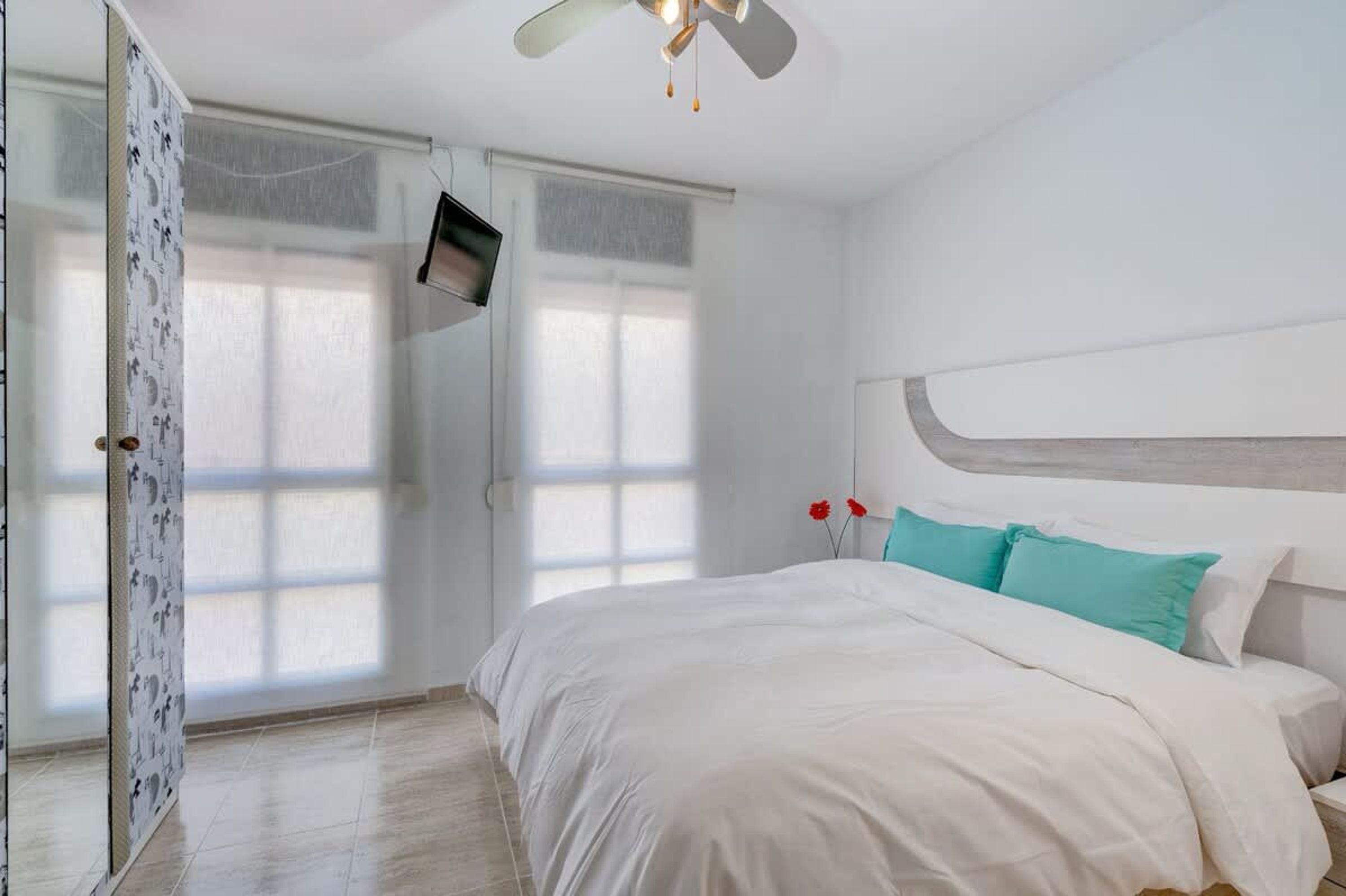 A bedroom with a bed, a ceiling fan, a TV mounted on the wall, and a window with curtains.