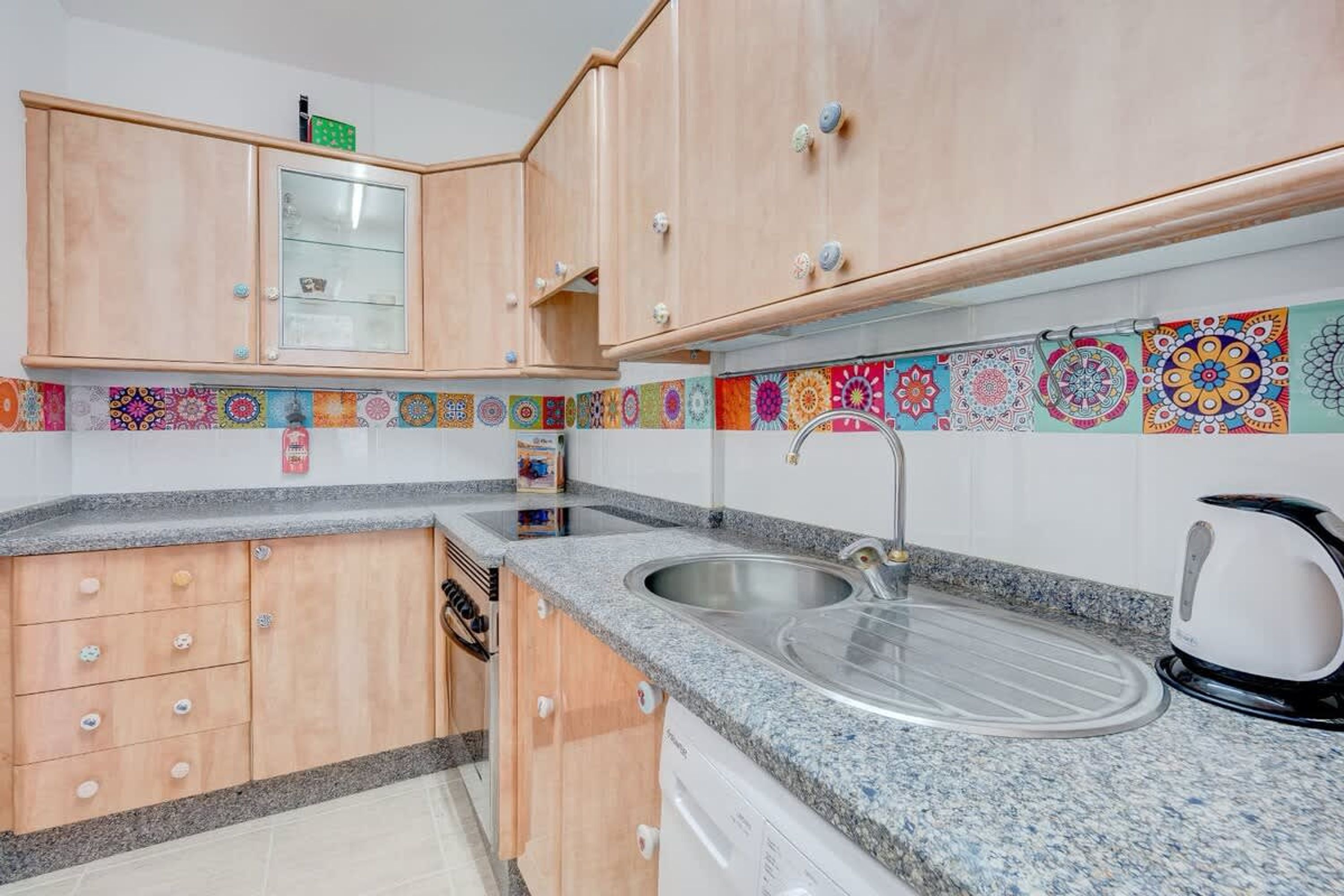 A kitchen with wooden cabinets, a granite countertop, a stainless steel sink, and a colorful tile backsplash.