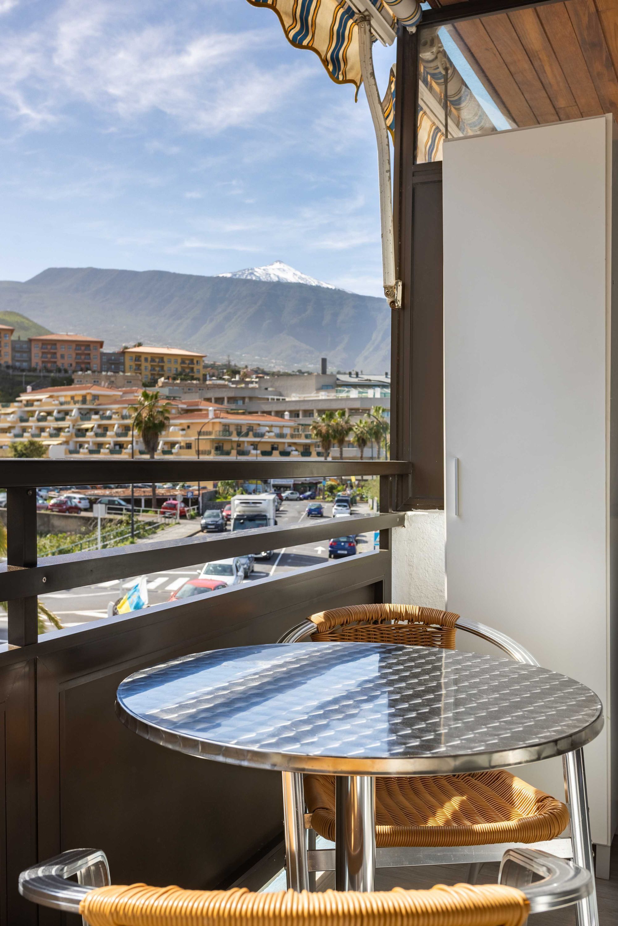 A balcony with a round glass table and wicker chairs, overlooking a parking lot and buildings with a mountain in the background.