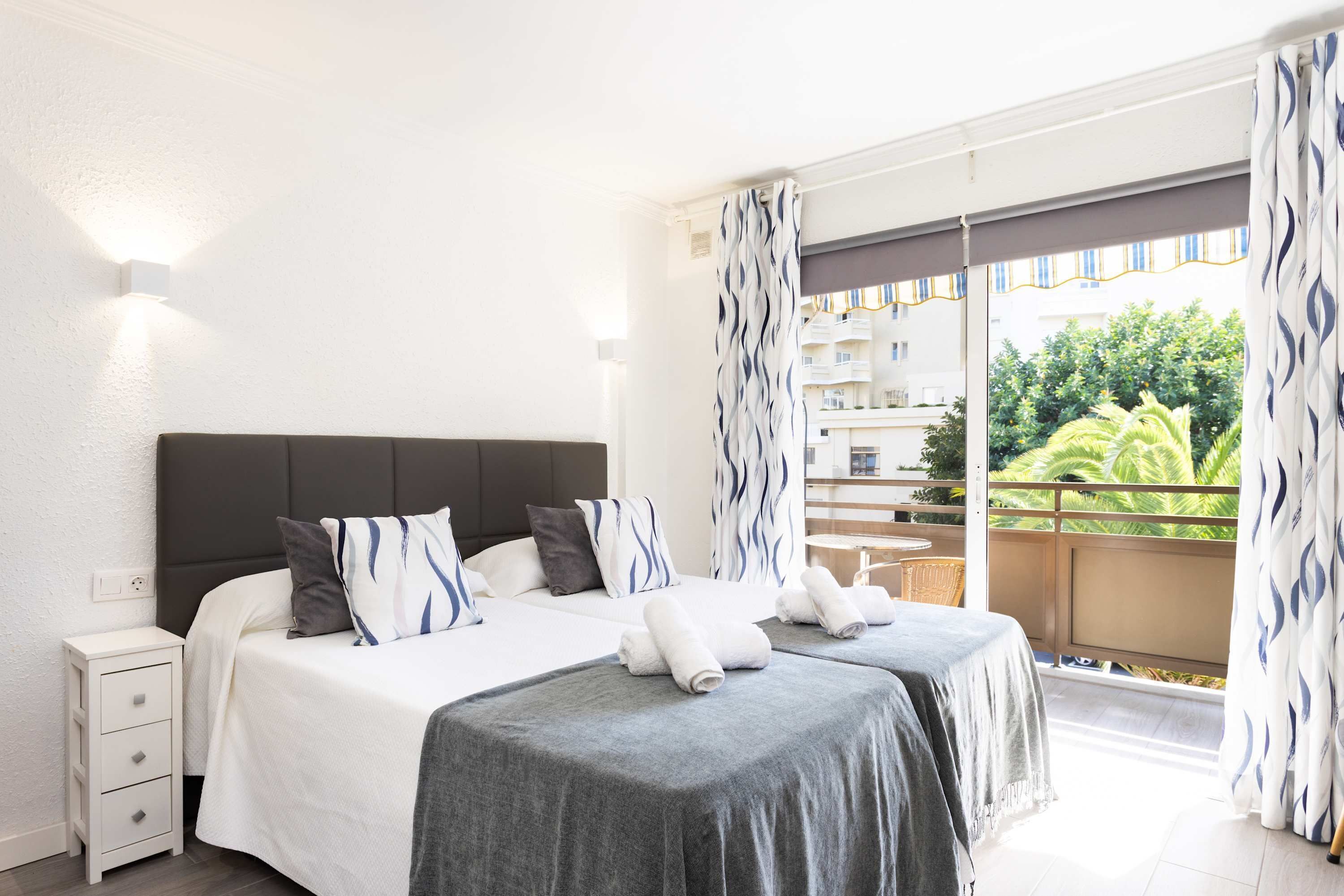A hotel room with a bed, a nightstand, a balcony with a view of buildings and greenery.