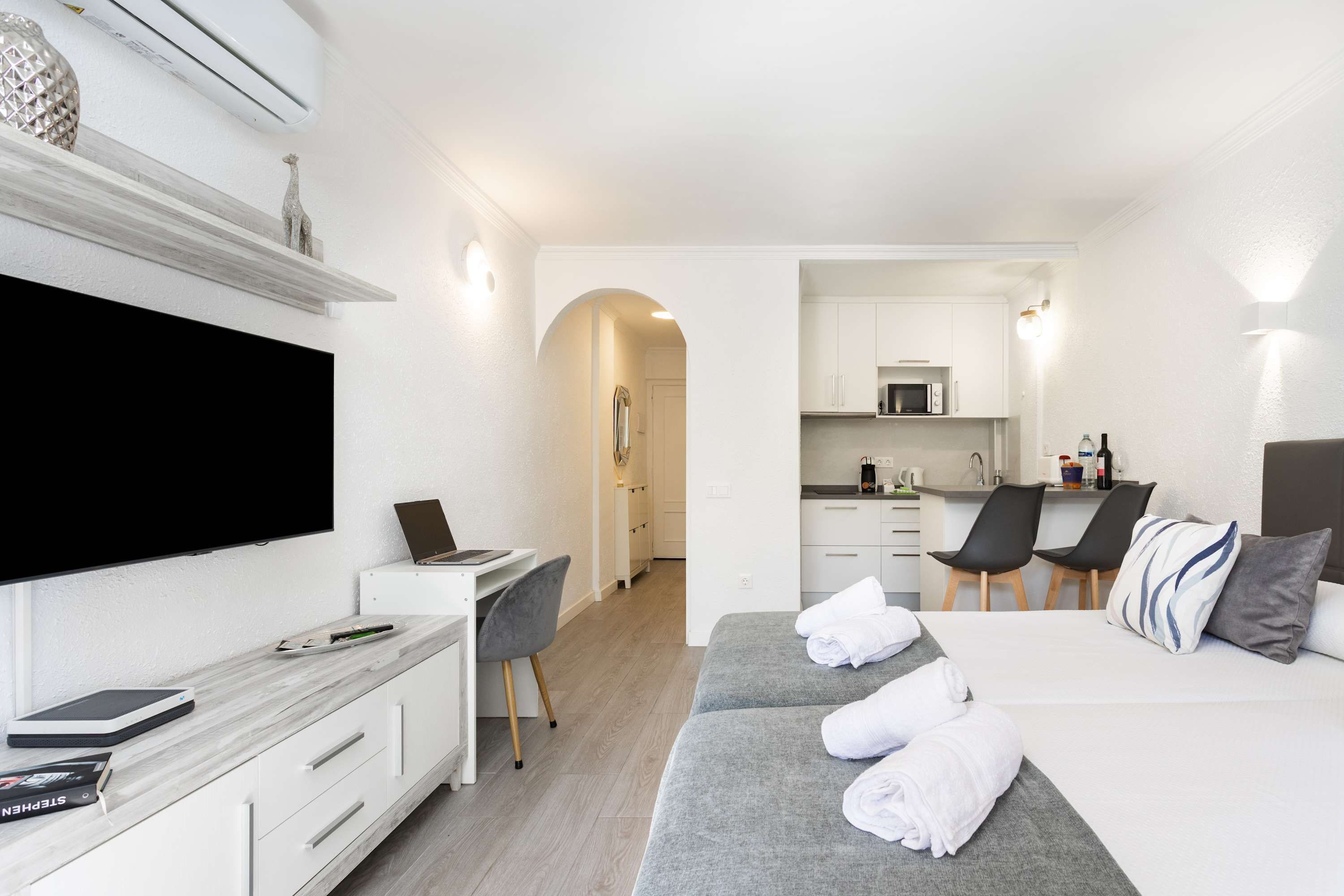 A modern hotel room with a flat-screen TV, a desk with a laptop, a bed with towels, a kitchenette, and a dining area.