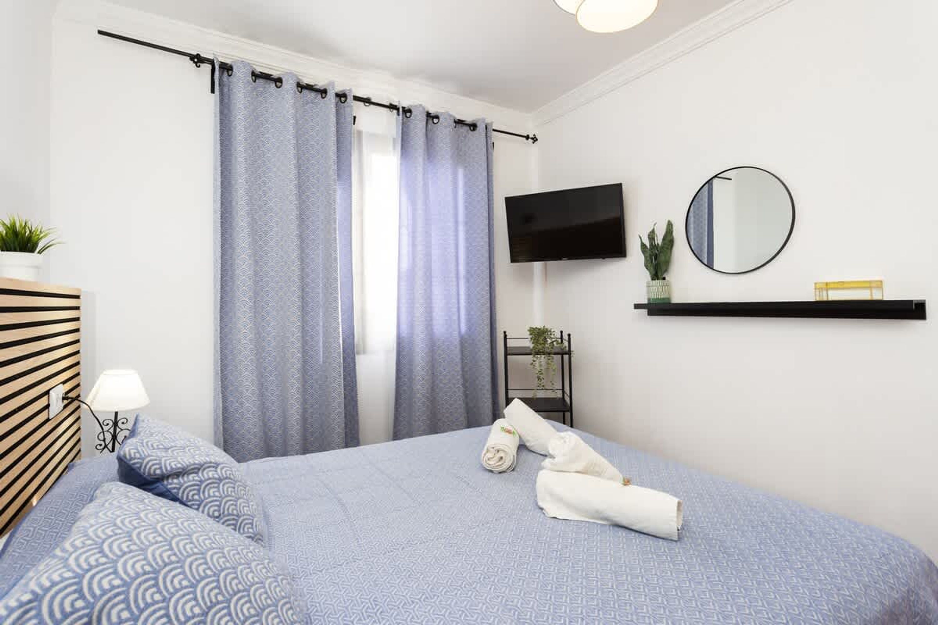 Een slaapkamer met een bed, gordijnen, een televisie, een spiegel en een plank met een plant.