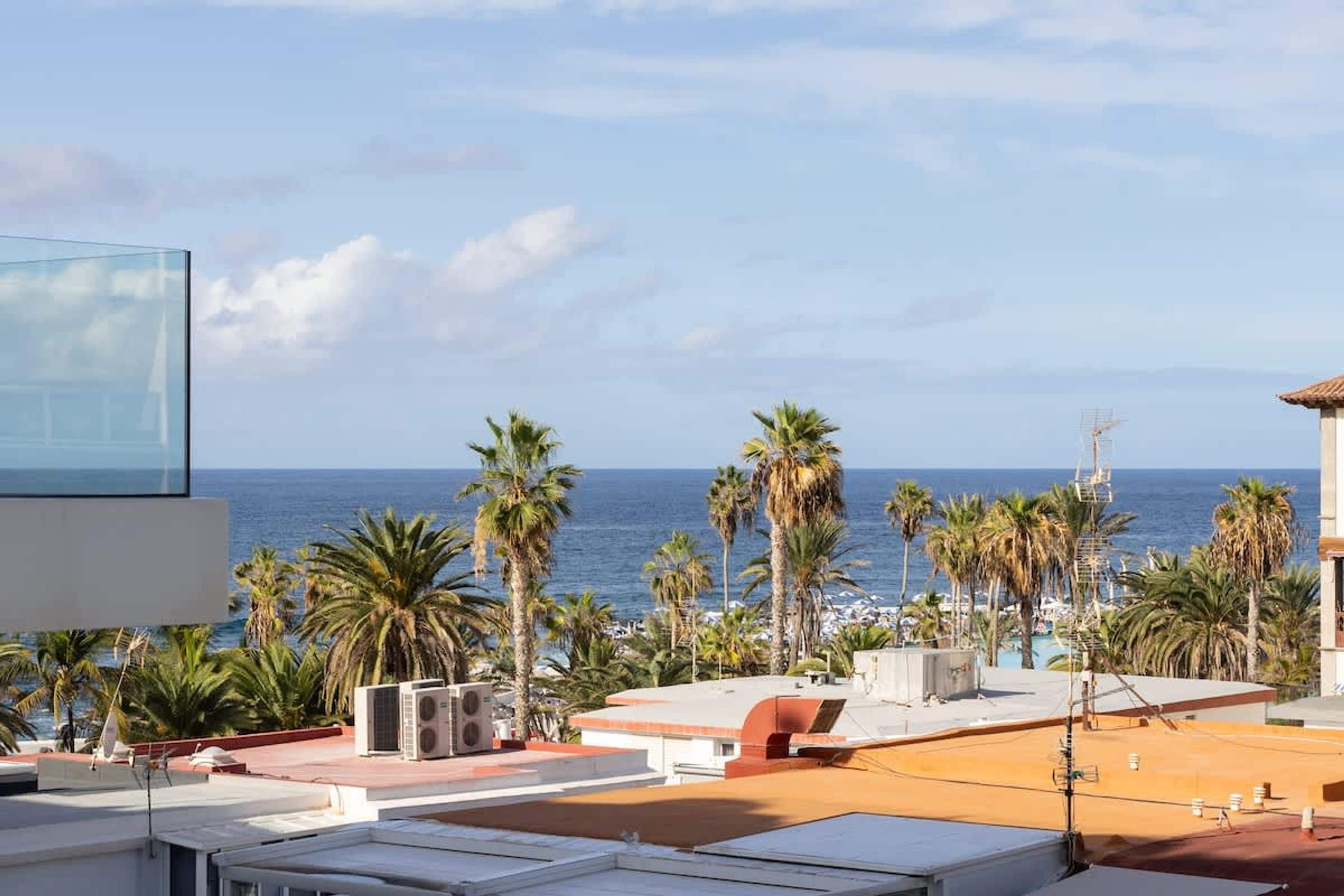 A rooftop terrace with a view of the sea, palm trees, and a clear sky.