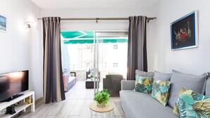 Een moderne woonkamer met een flatscreen-tv, een grijze bank met kussens met tropische patronen, een klein rond salontafeltje met een plant, en een schuifdeur die leidt naar een buitenruimte.