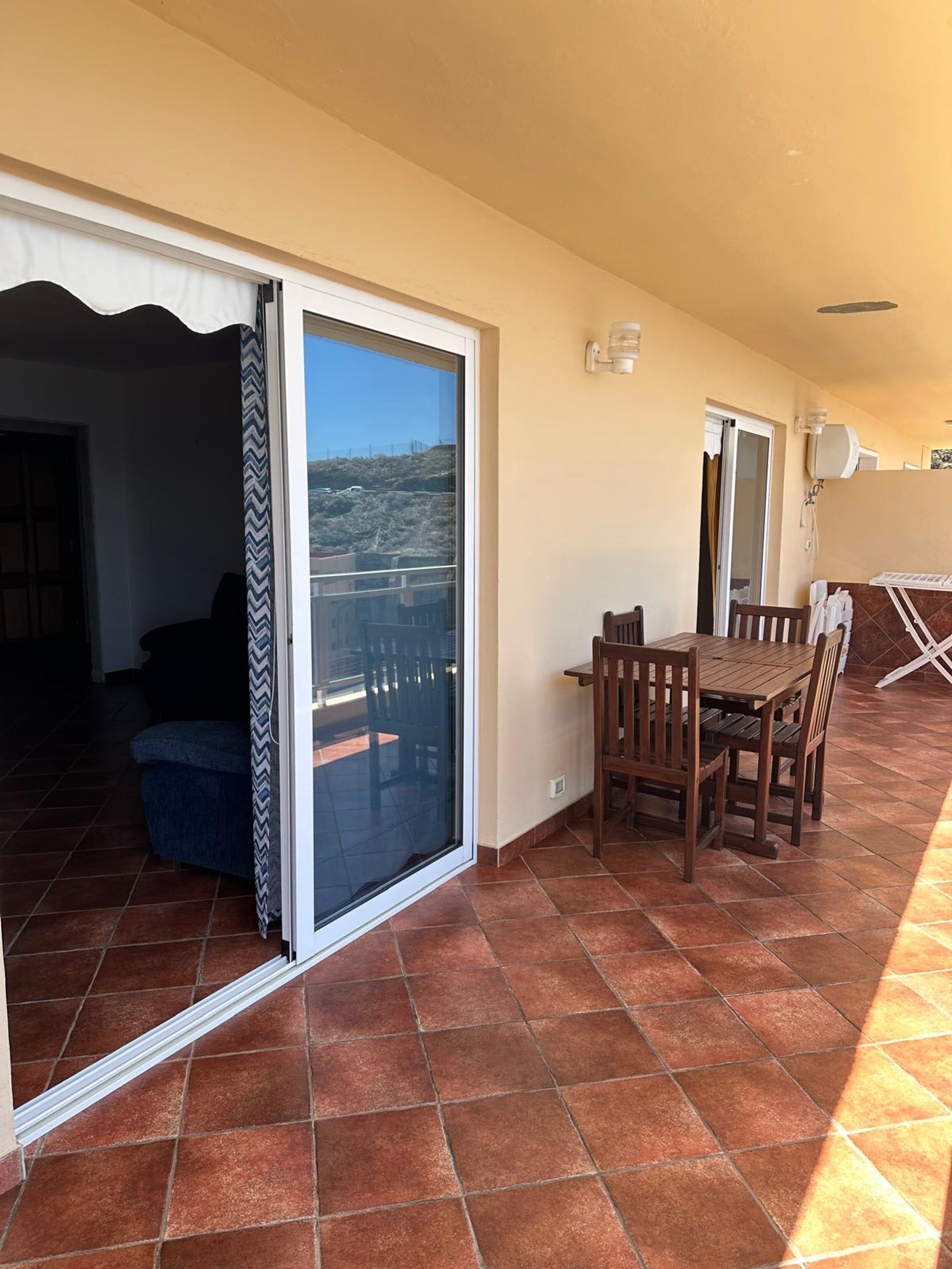 A balcony with a sliding glass door, a dining table with chairs, and a view of a hilly landscape.