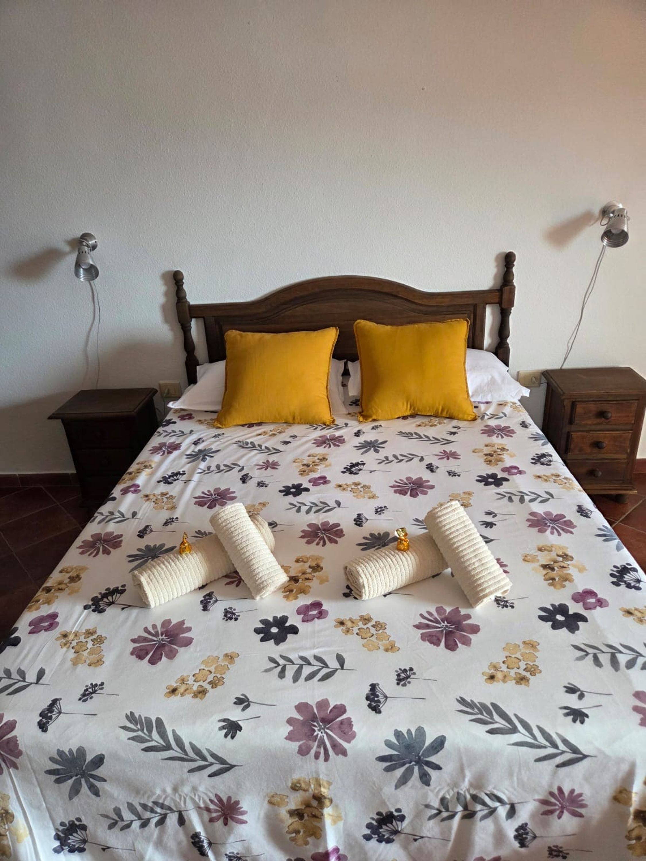 Una cama con colcha estampada, dos almohadas, cabecera de madera y mesita de noche con lámpara.