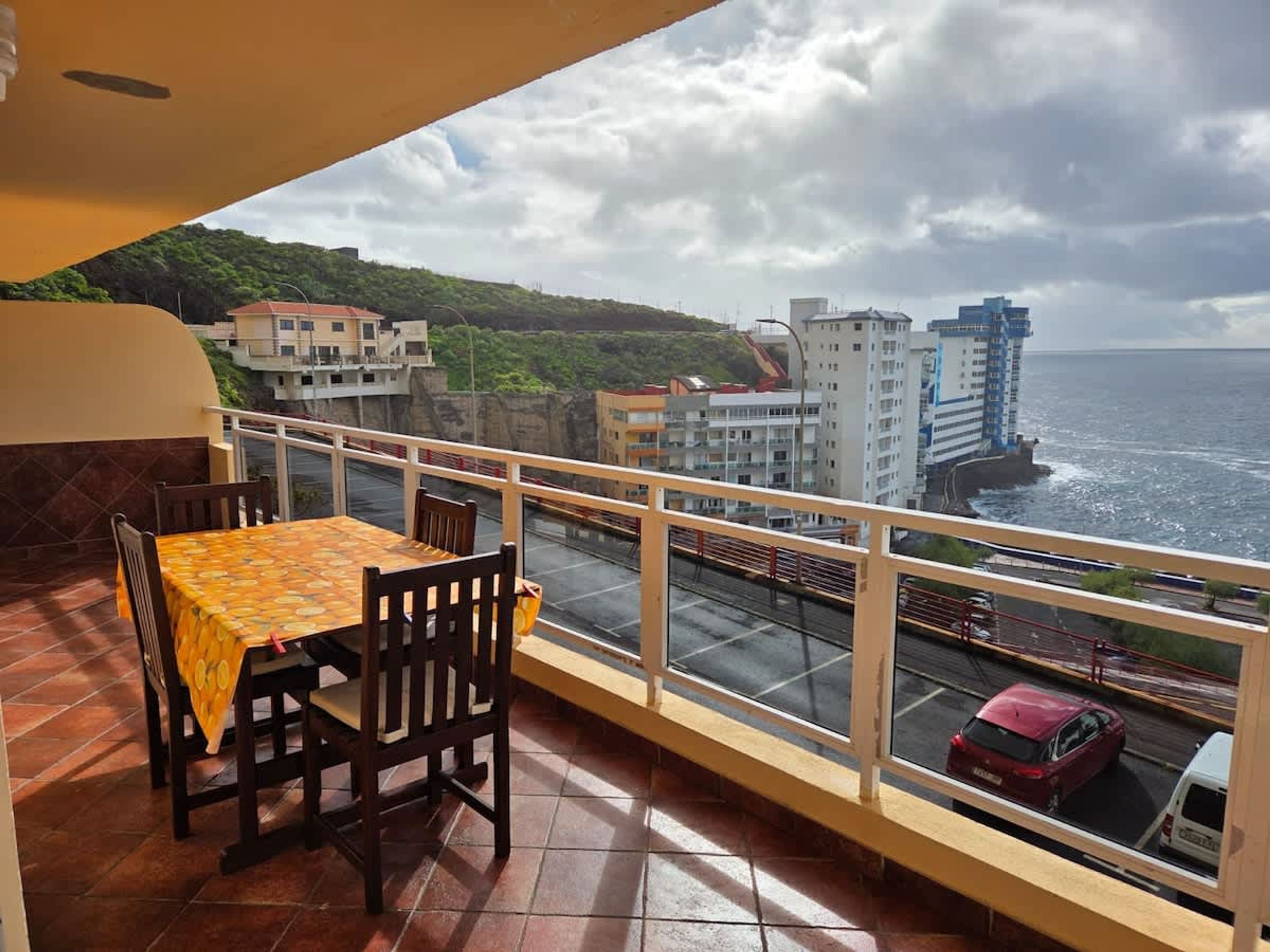 A balcony with a table and chairs, overlooking a coastal road with buildings and a cruise ship.