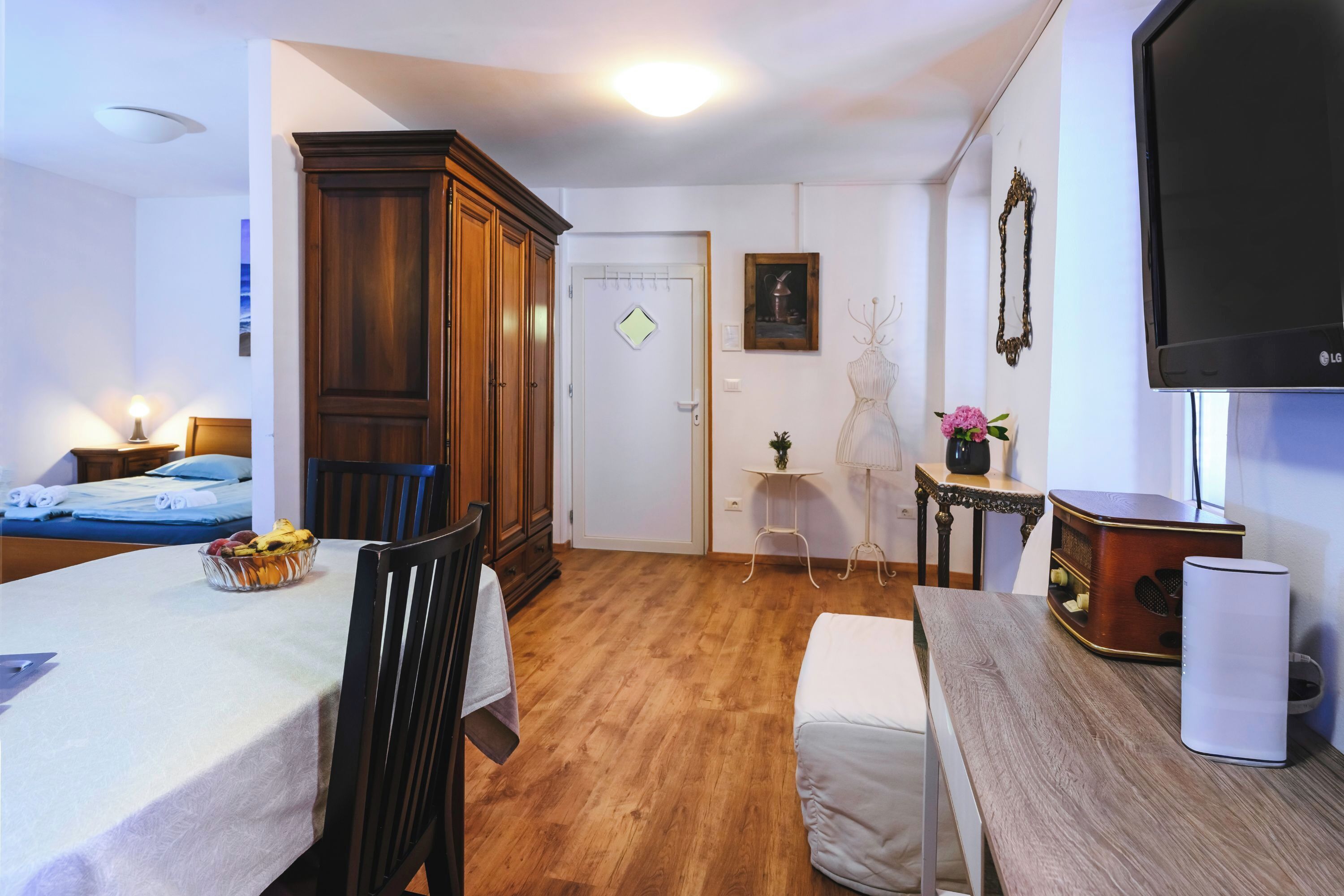 A room with a bed, a dining table with chairs, a TV, a wooden wardrobe, and a small table with a lamp and flowers.