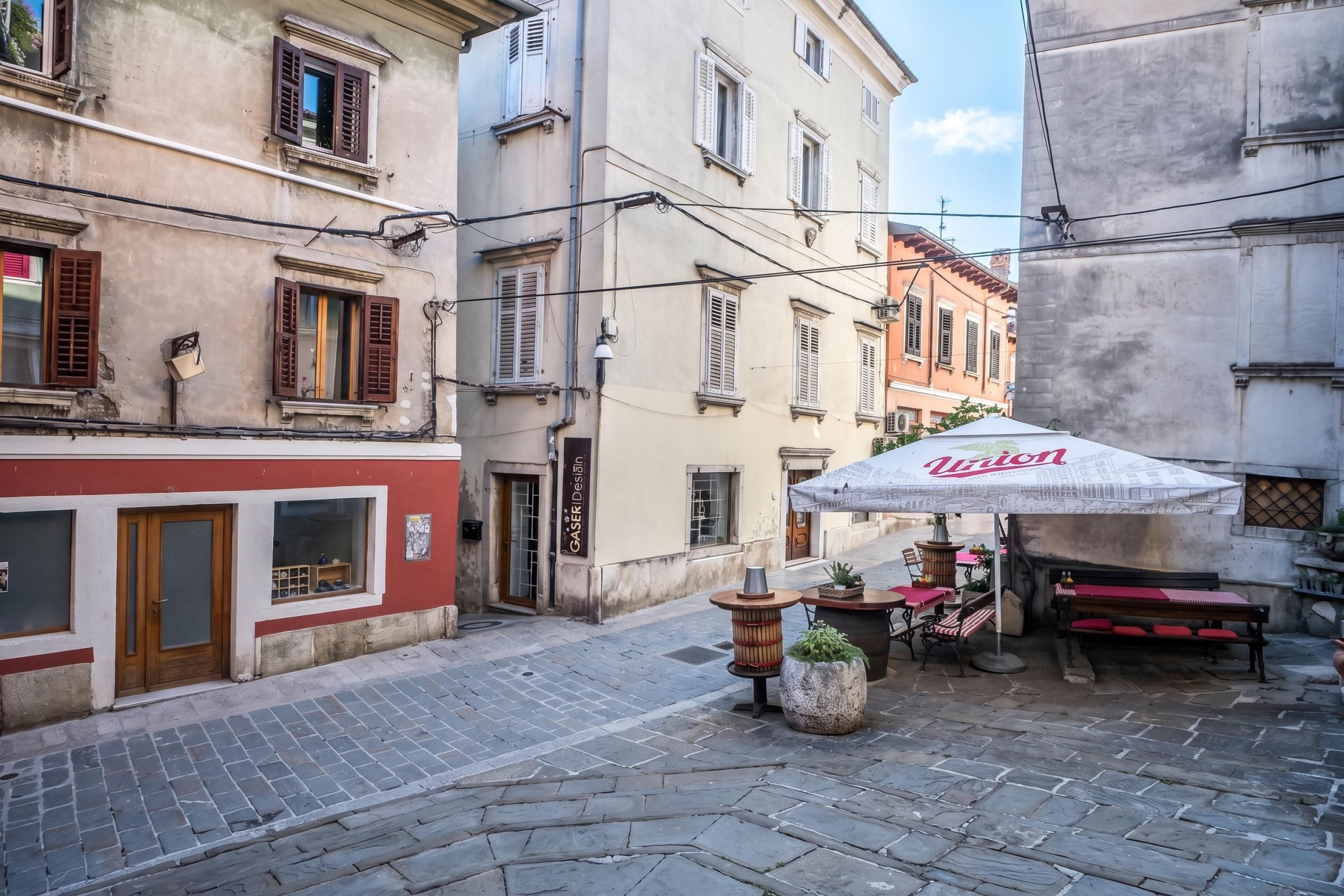A narrow street with cobblestone pavement, traditional European buildings with shuttered windows, and a café with an umbrella.
