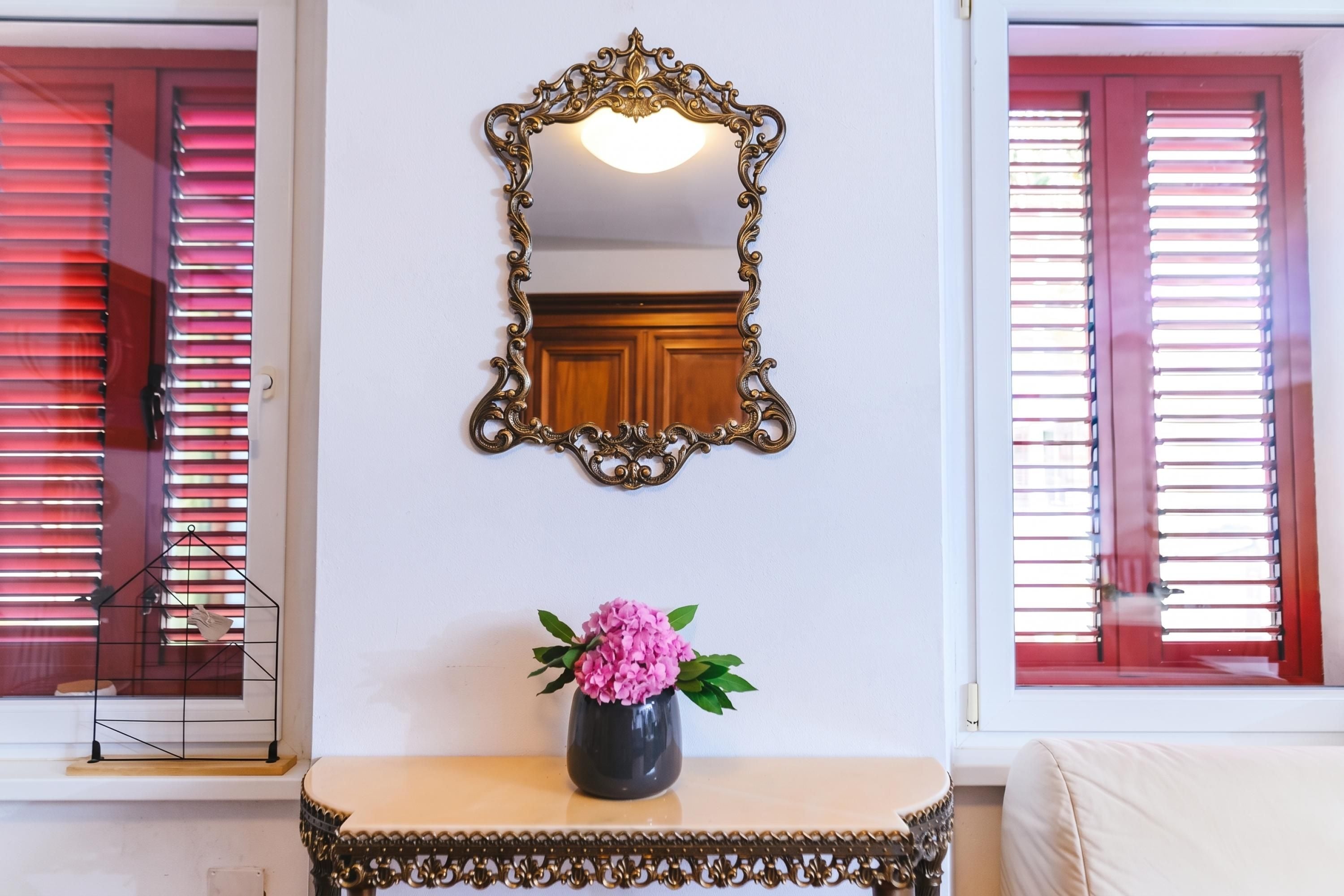 Une pièce agrémentée d’un miroir décoratif, d’une petite table sur laquelle est posé un vase de fleurs, et d’une fenêtre avec des stores.