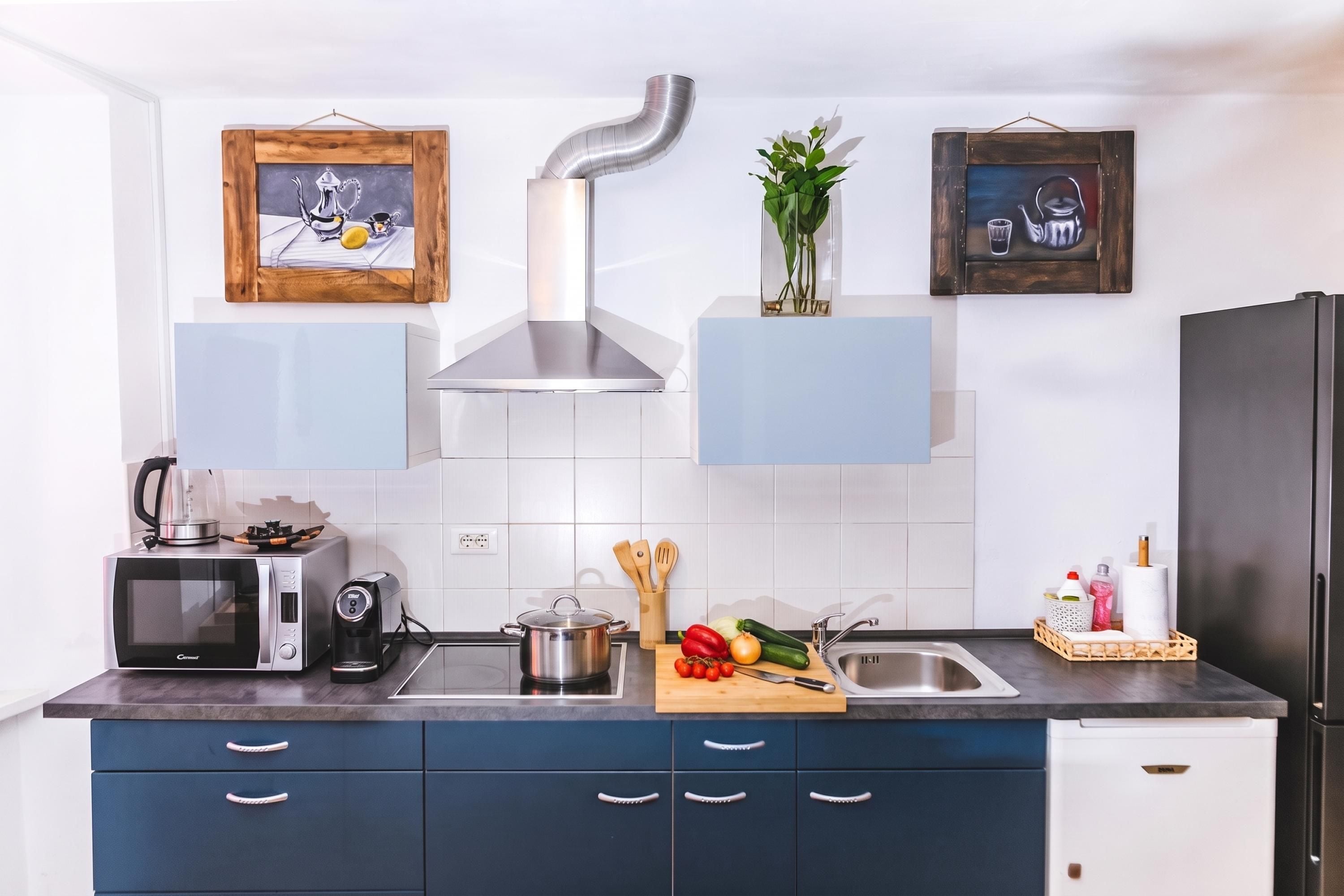 Eine moderne Küche mit Mikrowelle, Wasserkocher und einem Topf auf der Arbeitsfläche. Darunter befinden sich Holzauszieher und ein Kühlschrank rechts.