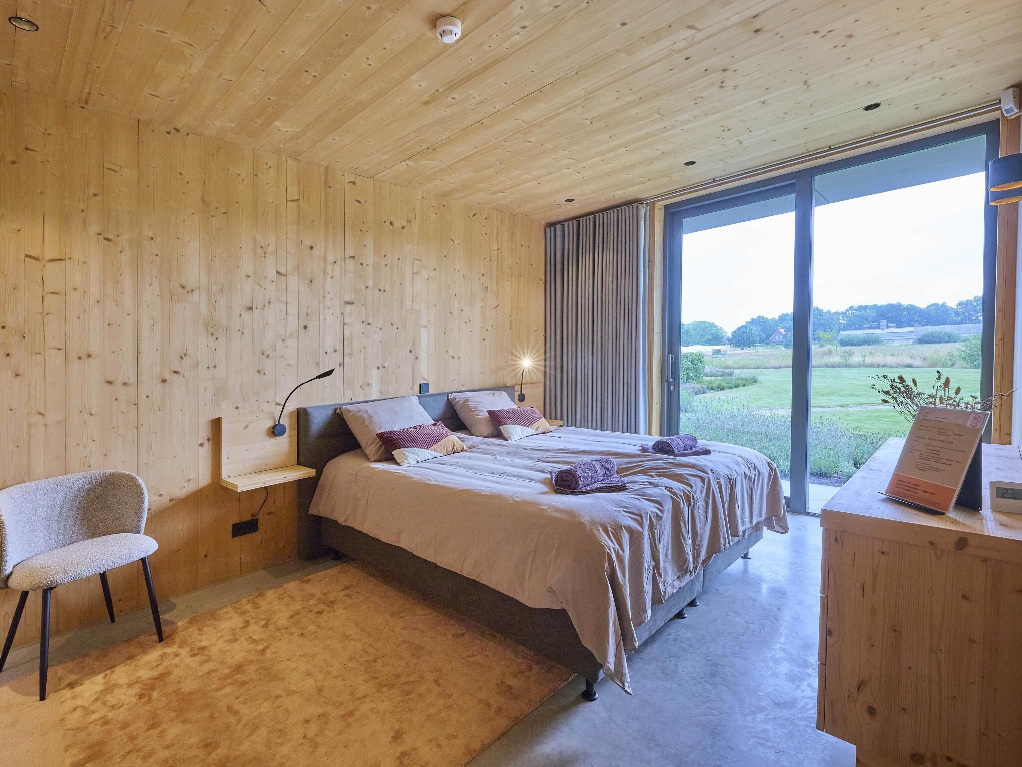 A bedroom with a large bed, a chair, a bedside table, and a view of the outdoors.