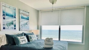 Una camera da letto con un letto, cuscini, un comodino, una lampada, una finestra vista mare e opere d'arte appese al muro.