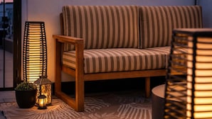 Et hyggeligt udendørs opholdsområde med en stribet sofa, en træstol og to høje gulvlamper.