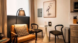 En moderne stue med en lædersofa, to lænestole, et rundt bord, en lampe, et indrammet kunstværk og et tekøkken.