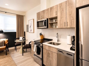 A compact kitchen with wooden cabinets, a built-in microwave, a stove, and a dishwasher. - Roami at Zuni | Studio Apartments | 2 Units (Denver)