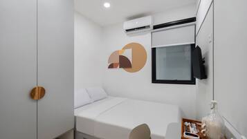 A modern hotel room with a bed, a desk, a chair, an air conditioning unit, and a television.