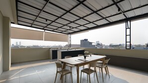 A rooftop terrace with a dining table, chairs, a small kitchen area, and a view of the cityscape.