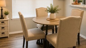 A dining area with a round table, four chairs, a window with blinds, a wall-mounted shelf with plants, and a kitchen area in the background. - 3-bedroom house in peaceful Houston with cool AC breeze (Houston)
