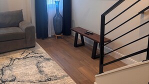 En stue med sofa, sofabord, fjernsyn monteret på væggen, trappe og et stort vindue med gardiner.