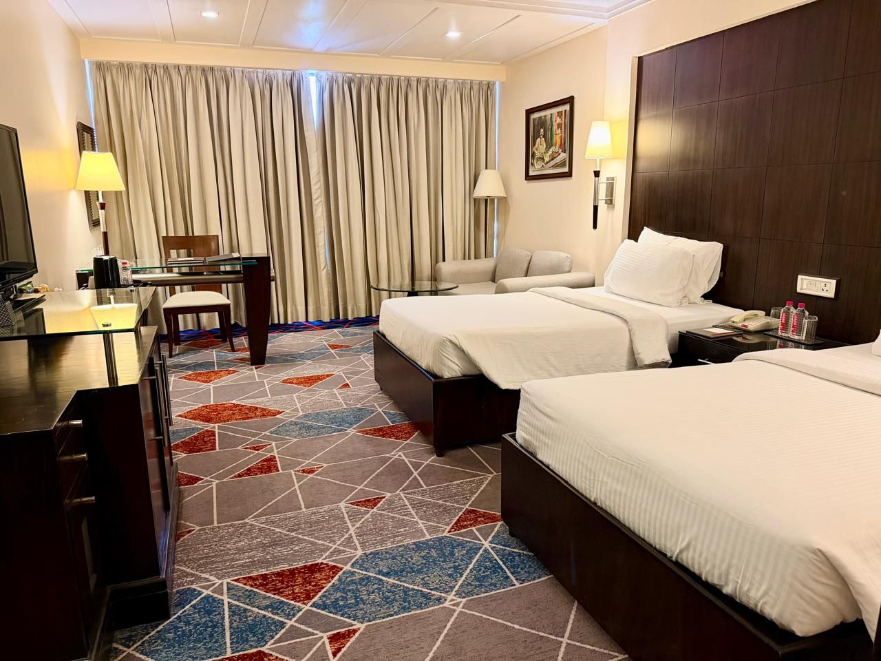 A hotel room with two beds, a desk, a chair, a TV, and a patterned carpet.