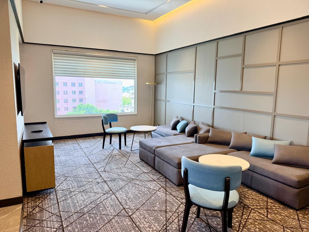 A modern hotel room with a sectional sofa, a small round table, a desk, and a large window with blinds.