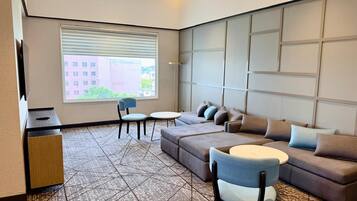 A modern hotel room with a sectional sofa, a small round table, a desk, and a large window with blinds.