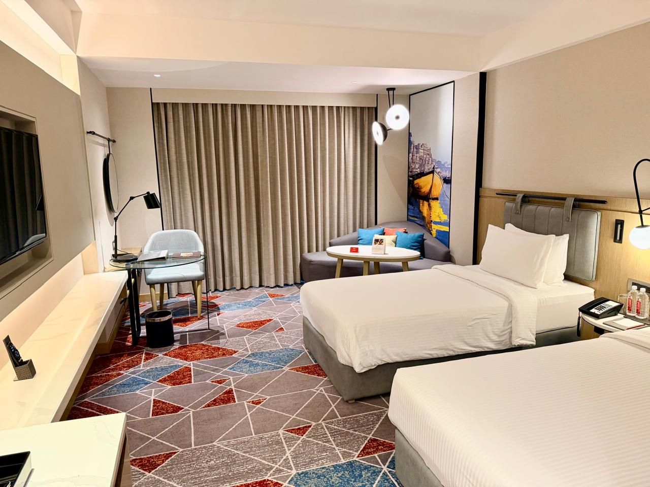 Ein Hotelzimmer mit zwei Betten, einem Schreibtisch, einem Sessel, einem kleinen Tisch und einem großen Fenster mit Vorhängen.
