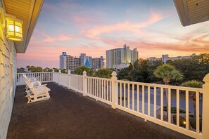 A balcony with a white railing, a row of white lounge chairs, and a view of a city skyline at sunset. - Stunning Oasis ~ Pool ~ Hot Tub ~ Elevator ~ Poker ~ Spacious Floorplan ~ Tiki (North Myrtle Beach)