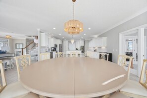A spacious dining area with a round table and chairs, a chandelier, and a kitchen in the background.