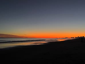 Ein Strand bei Sonnenuntergang mit klarem Himmel, ruhiger See und einer fernen Stadtansicht.