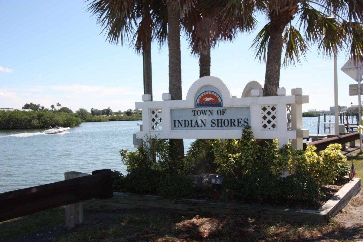 A sign for the Town of Indian Shores with a view of the water and palm trees.