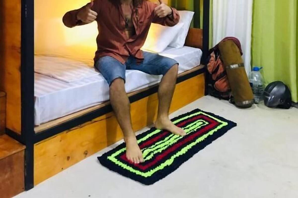Quarto com beliche, uma pessoa sentada na cama inferior, um travesseiro, um cobertor e um tapete no chão.