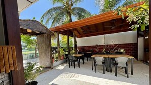 Une terrasse extérieure avec des tables et des chaises, une structure à toit de chaume et des palmiers en arrière-plan.