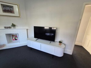 Ein Wohnzimmer mit Flachbildfernseher auf einer weißen Konsole, einem Kamin mit einem darüber hängenden Bild und einer kleinen Topfpflanze auf der Konsole.