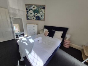 A neatly made bed with a white comforter and pillows, a bedside lamp, and a floral painting on the wall. - Comfortable Apartment in Fremantle (Fremantle)