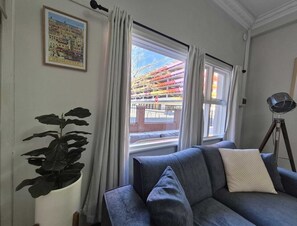 Ein Zimmer mit einer Couch, einem Blumentopf, einem gerahmten Bild und einem Fenster mit Blick auf ein Gebäude.