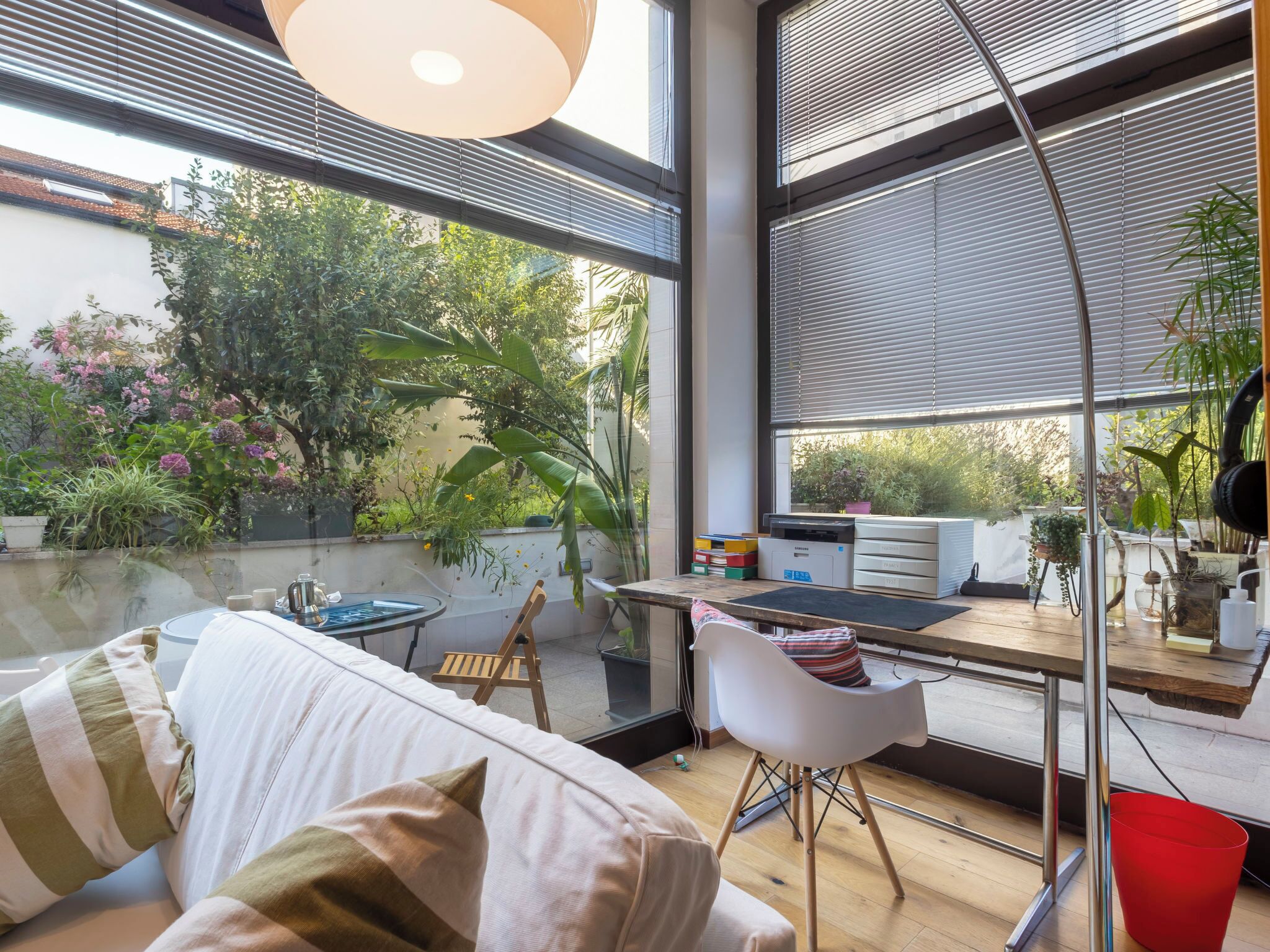 A modern living space with a sofa, a dining table, and a chair. There is a large window with blinds, and a view of greenery outside.
