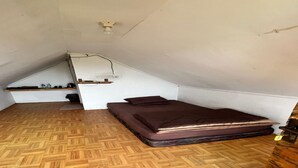 A small, triangular attic bedroom with a wooden floor, a single bed, a shelf with items, and a ceiling fan.
