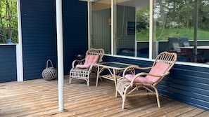 Eine Holzterrasse mit Korbmöbeln und einer Glastür, die zu einem überdachten Bereich führt.