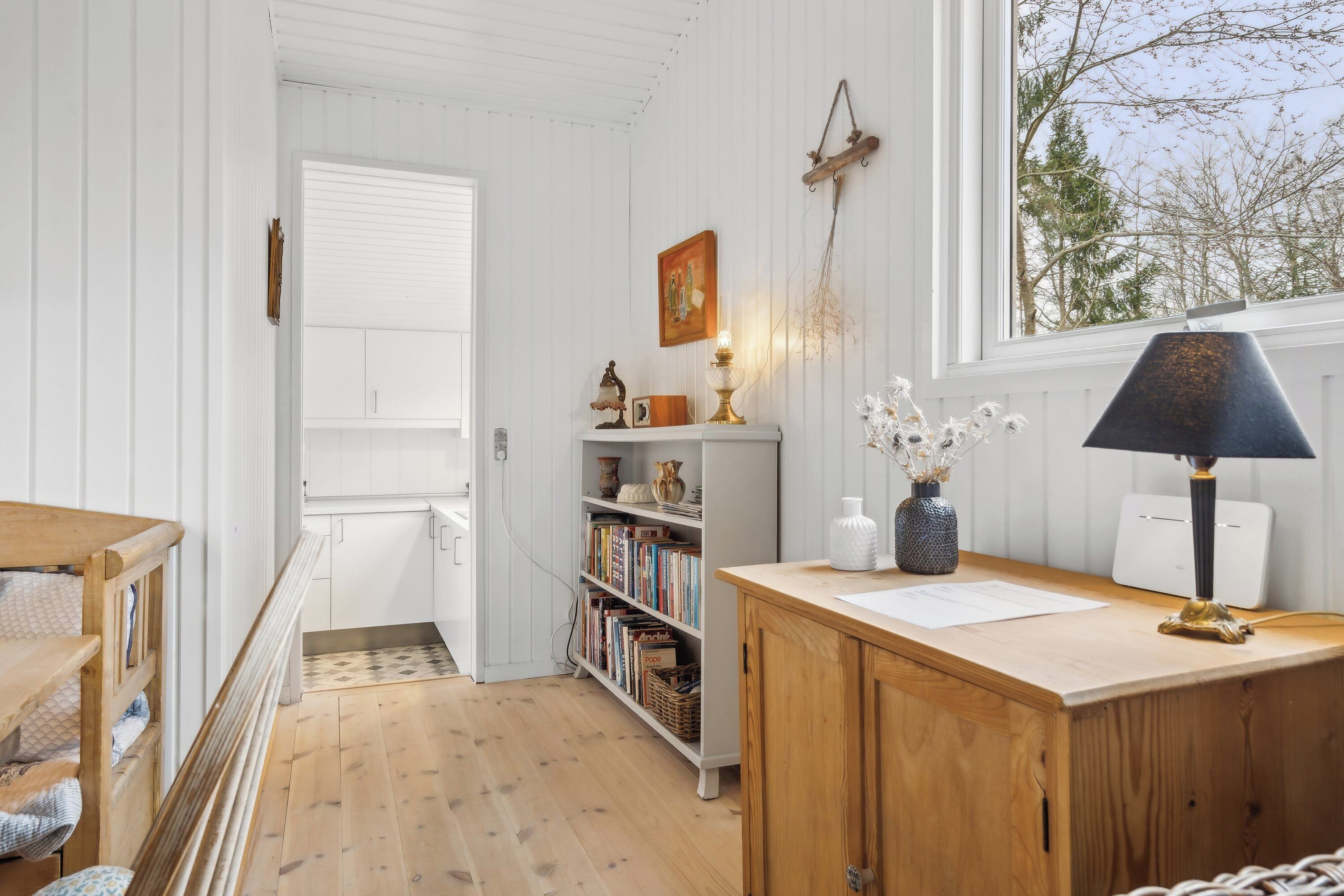 A room with a wooden desk, a bookshelf, and a window with a view of trees.
