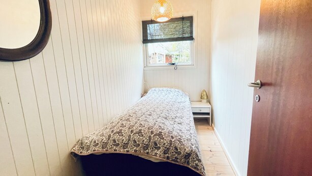Une petite pièce comprenant un lit, une fenêtre avec un store, une petite table avec une lampe et une porte.