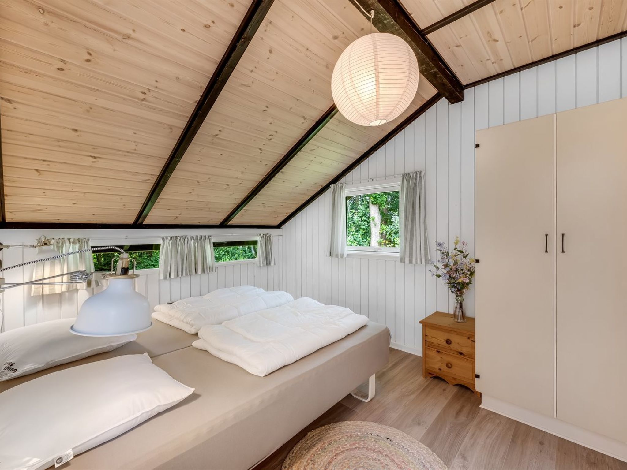 Een slaapkamer met een houten plafond, een bed met witte beddengoed, een nachtkastje met een lamp, een kledingkast en een raam met gordijnen.