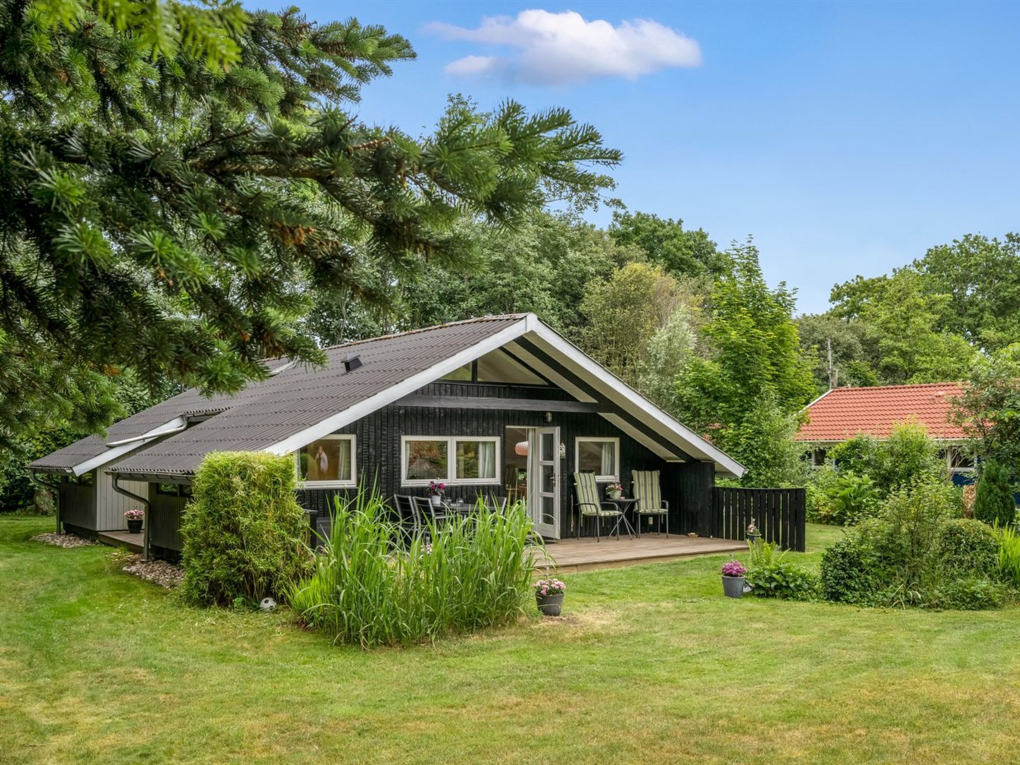 Une petite cabane aux couleurs sombres, dotée d’un toit en pente, d’un porche à l’avant avec deux chaises et d’un jardin bien entretenu.